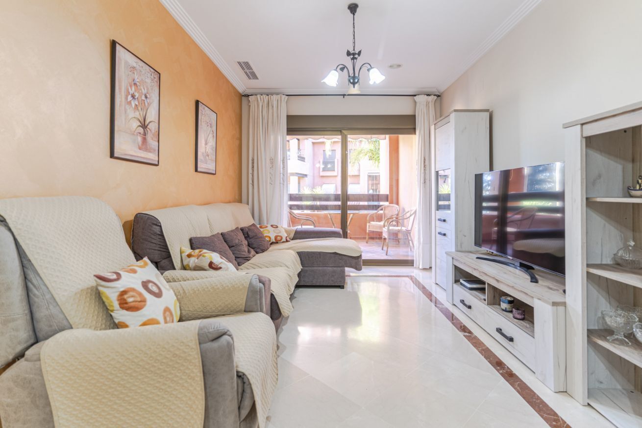 A living room with a sofa set, a TV on a wooden stand, and a balcony with chairs.