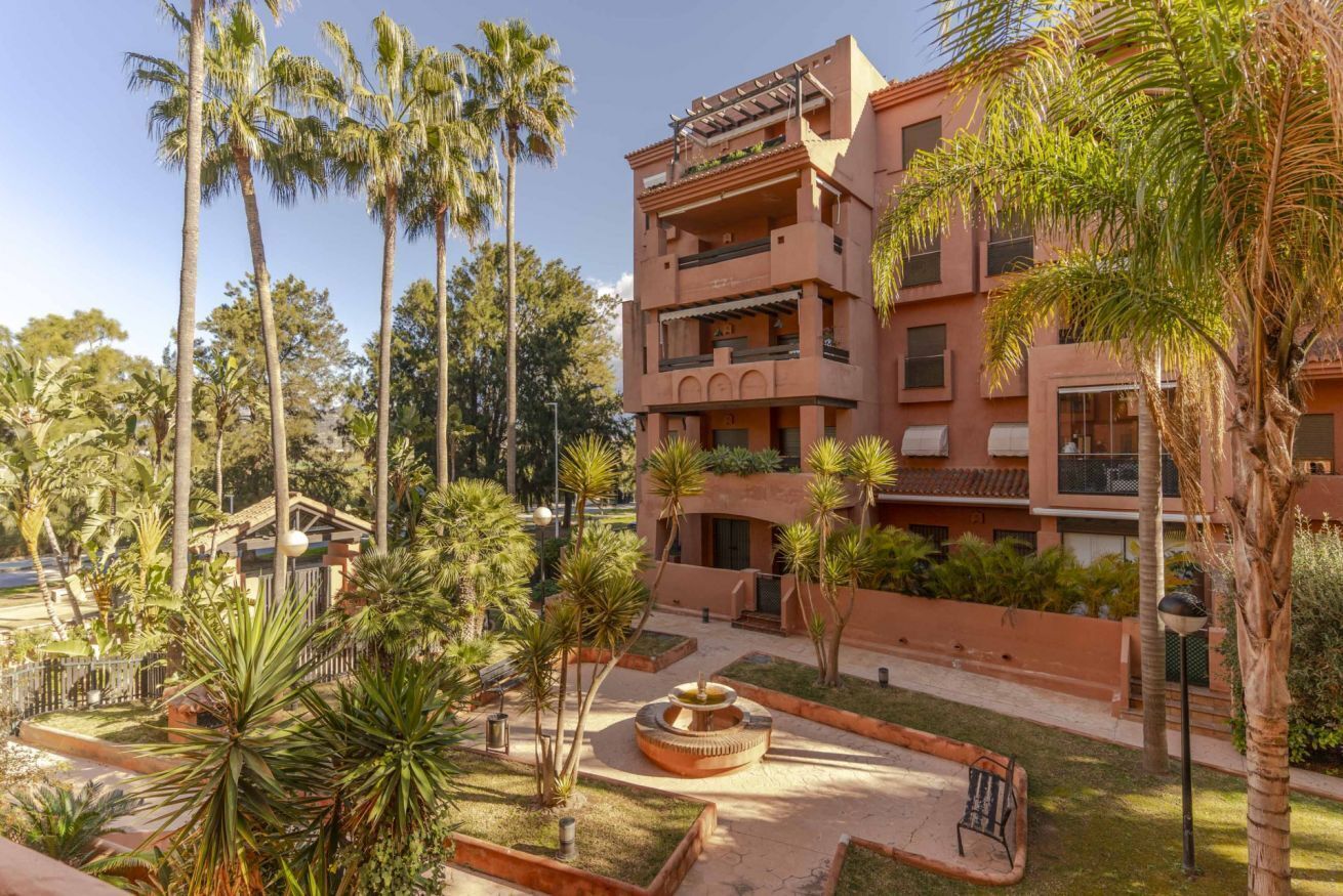 A multi-story building with balconies, surrounded by palm trees and a central courtyard with a fountain.