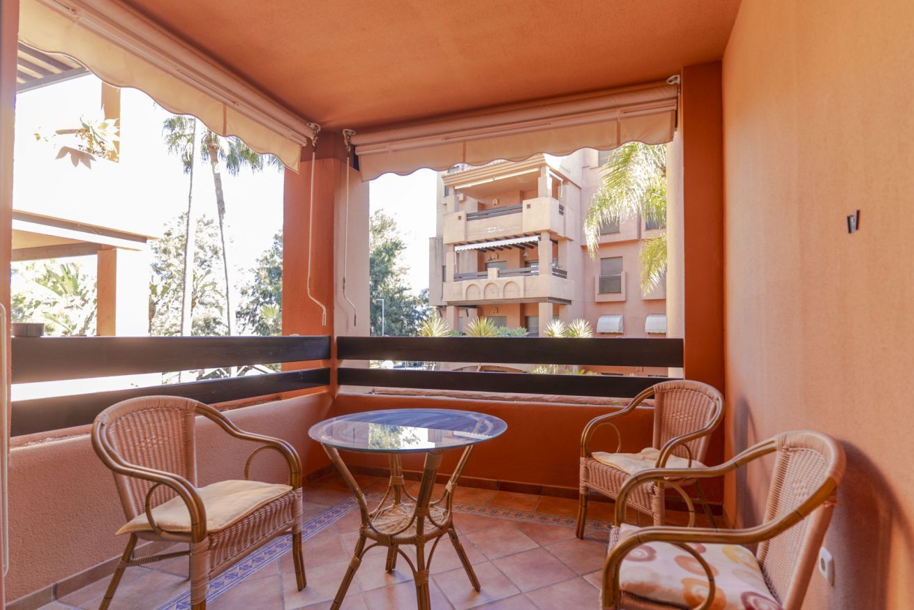 A balcony with wicker chairs and a glass table, overlooking a building with balconies and palm trees.