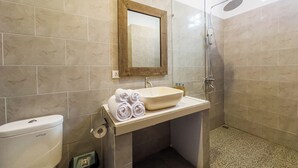 Une salle de bain avec un mur recouvert de carreaux beiges, un miroir à cadre en bois, un lavabo avec un receveur beige, des toilettes et une douche avec une porte en verre.