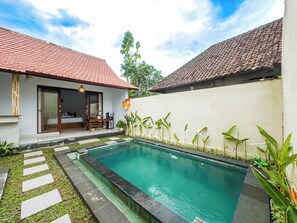 A swimming pool with a tiled deck, a wooden deck area with a table and chairs, and a white wall with a red-tiled roof. - Felicia House (Ubud)