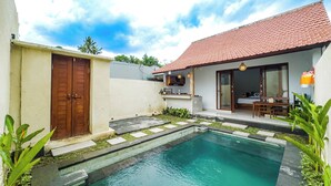 Une piscine avec une terrasse carrelée, une porte en bois et un petit jardin agrémenté de plantes.