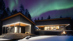 Et moderne hus med en overdækket veranda og en pejs, omgivet af sne og under en nattehimmel med nordlys.