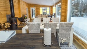 Interior acogedor de una cabaña con una mesa de comedor central, sillas de mimbre, una chimenea y ventanas amplias que ofrecen vistas de un paisaje nevado.