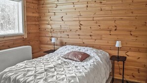Un dormitorio con pared de madera, una cama con edredón blanco, dos mesitas de noche con lámparas y una ventana con persianas.