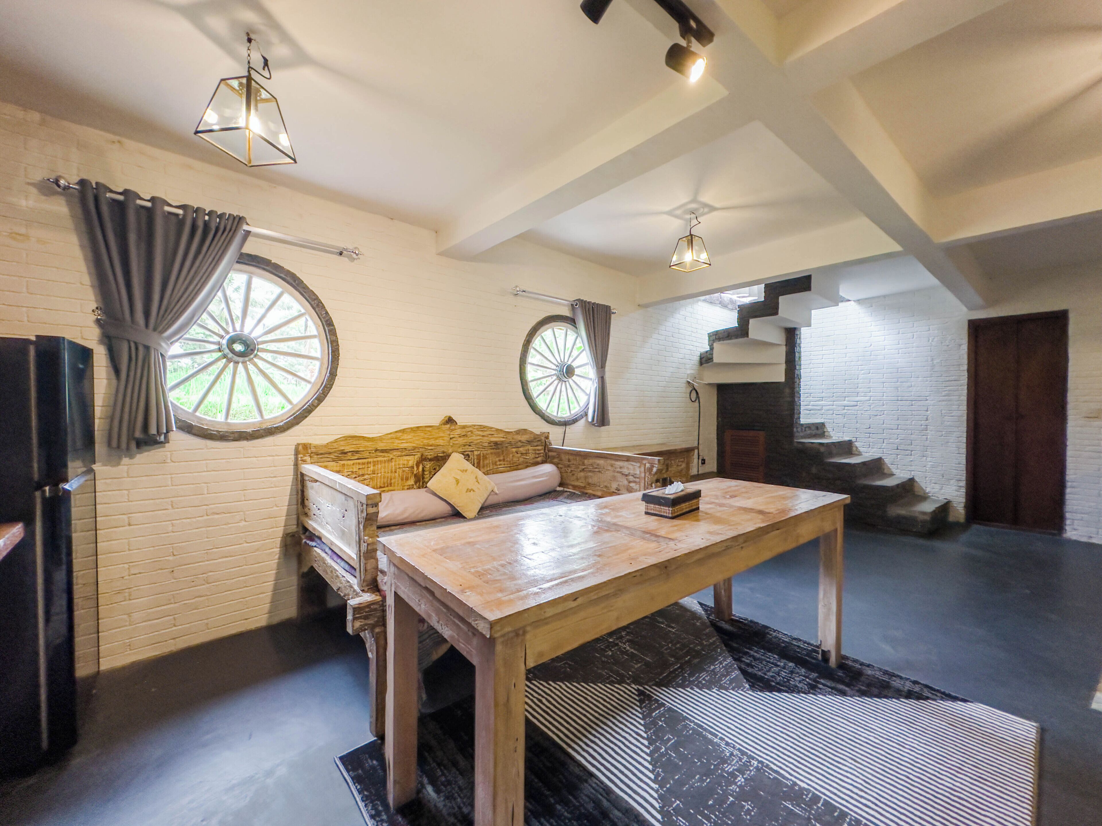 A spacious room with a large wooden table, two windows with curtains, a staircase, and a brick wall.