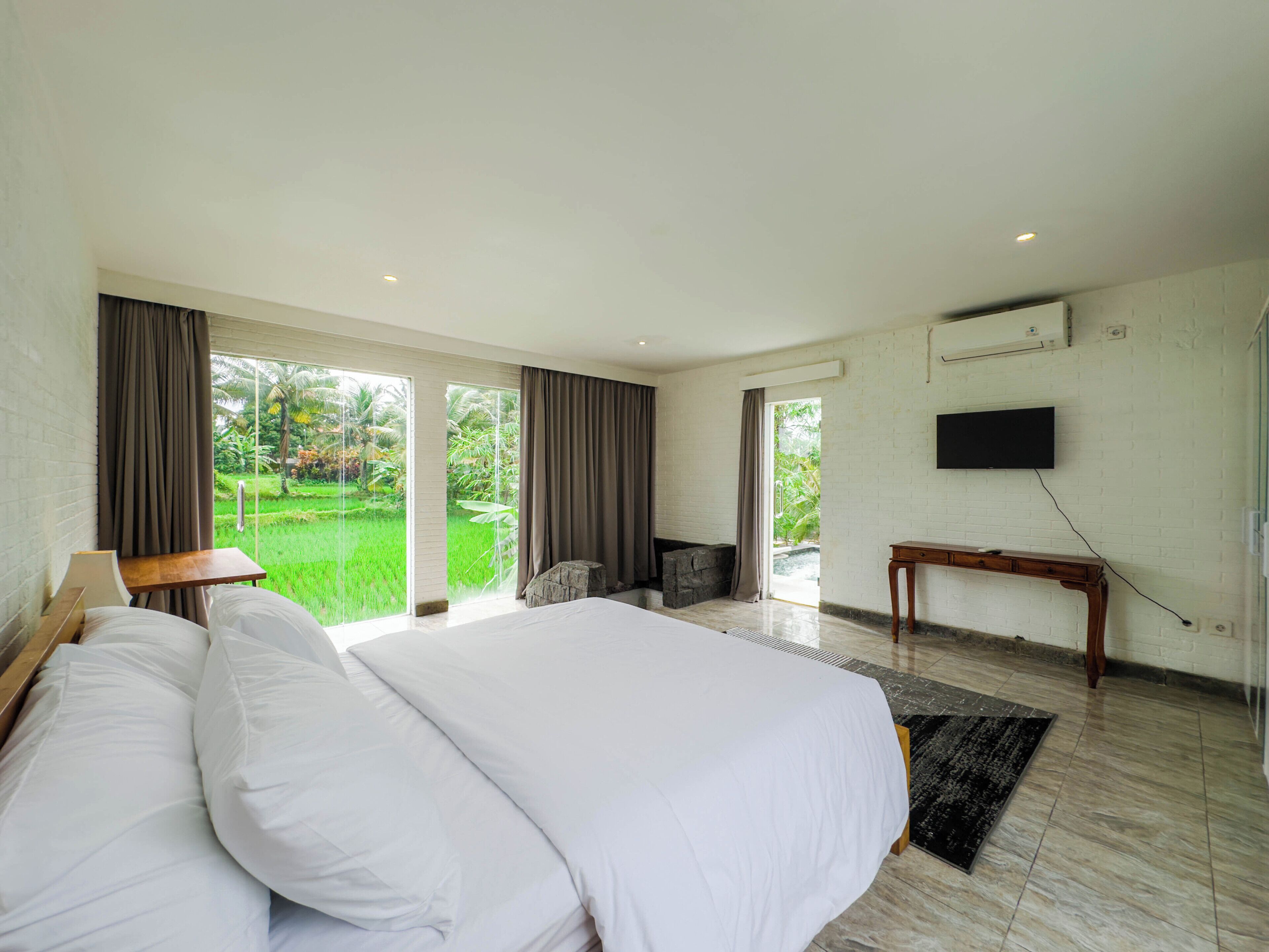 A bedroom with a large bed, a TV mounted on the wall, and a view of a lush garden through the window.