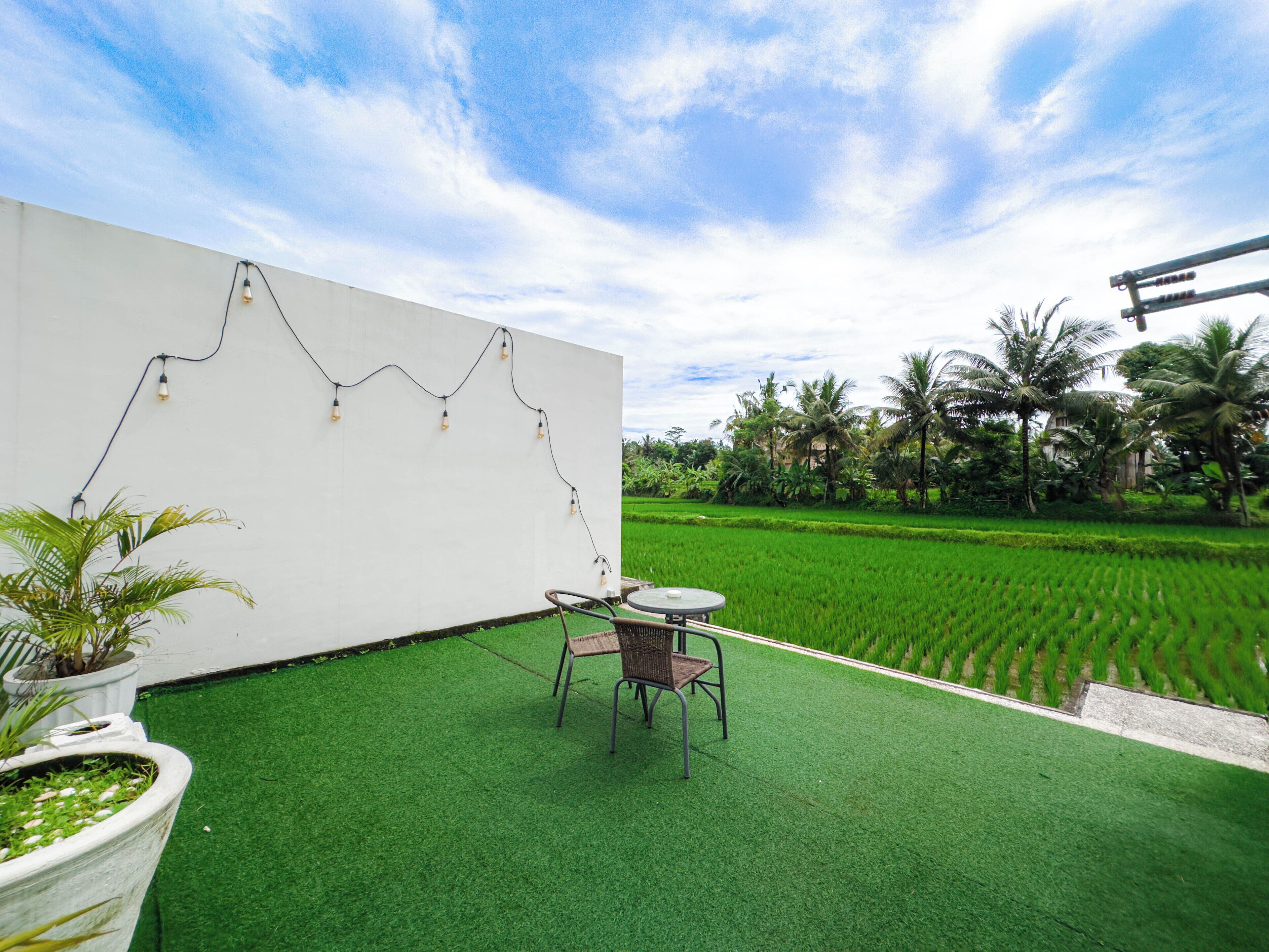 A modern outdoor seating area with a white wall, string lights, and a view of a lush green field.