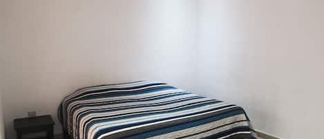 Una habitación pequeña con cama individual, colcha a rayas, mesita de noche negra y aire acondicionado de pared.