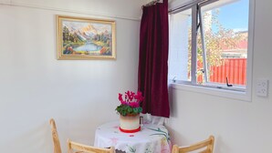 Zona pranzo con tavolo apparecchiato per due, un dipinto paesaggistico incorniciato al muro e una finestra vista su una recinzione rossa e vegetazione.