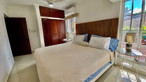 A bedroom with a large bed, wooden headboard, a small bedside table with a lamp, a window with blinds, and a door. - Apartamento Cerca Malecón by RHV Living (Puerto Plata)