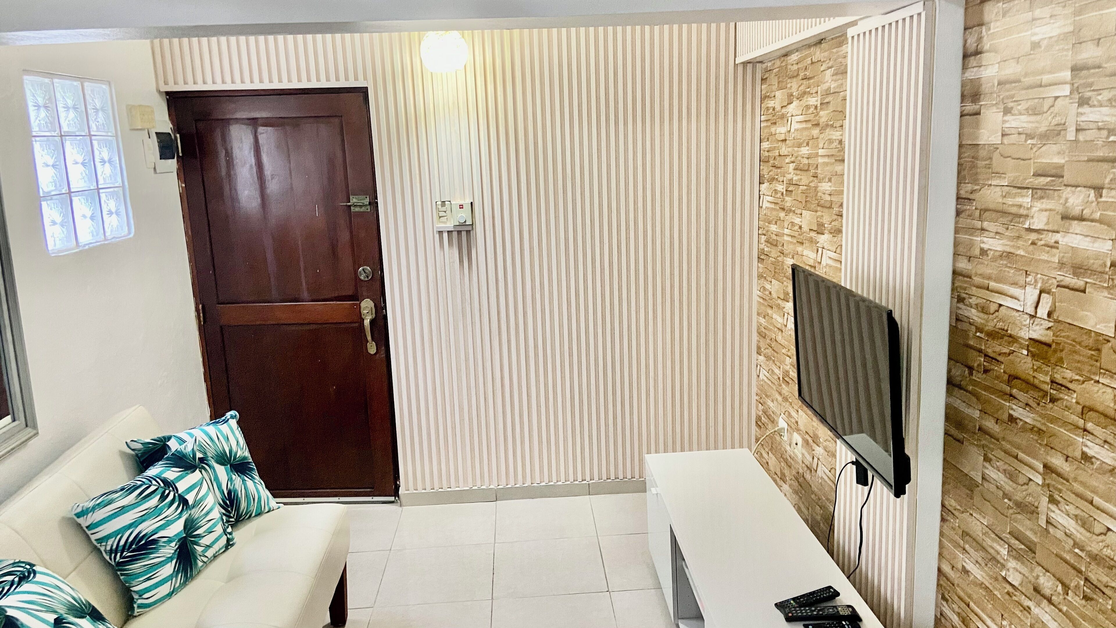 A living room with a wooden door, a white sofa with patterned cushions, a white coffee table, and a TV mounted on a stone-textured wall.