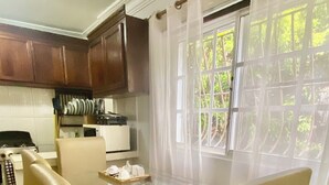 A dining area with a glass table and four chairs, a kitchen in the background, and a window with curtains.
