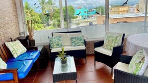 A covered outdoor seating area with wicker furniture, a glass table, and a view of the cityscape.