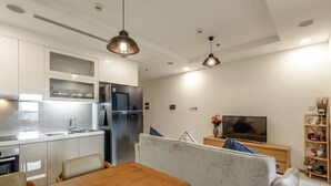 A modern kitchen with white cabinets, a built-in oven, and a stainless steel refrigerator. There is a dining area with a wooden table and chairs, and a grey sofa in the living room area.