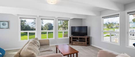 A living room with a sofa, a coffee table, and a TV on a wooden stand. There are large windows with a view of greenery and a building.