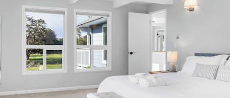 A bedroom with a large bed, a bench, and a view of a house through a window.