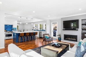 A modern living room with a fireplace, a TV mounted on the wall, and a kitchen area with blue tiles and a bar. - Spacious House in Walking Distance to the Beach (Aptos)