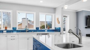 Une cuisine moderne avec des meubles blancs, des touches de bleu, un grand îlot central doté d’un évier et une vue sur l’extérieur grâce à de grandes fenêtres.