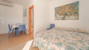Un dormitorio con cama, escritorio y sillas, un cuadro en la pared y una puerta que da a otra habitación.