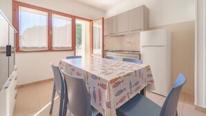 Een kleine keuken met een koelkast, een spoelbak en kastjes. Er is een eettafel met stoelen en een raam met gordijnen.