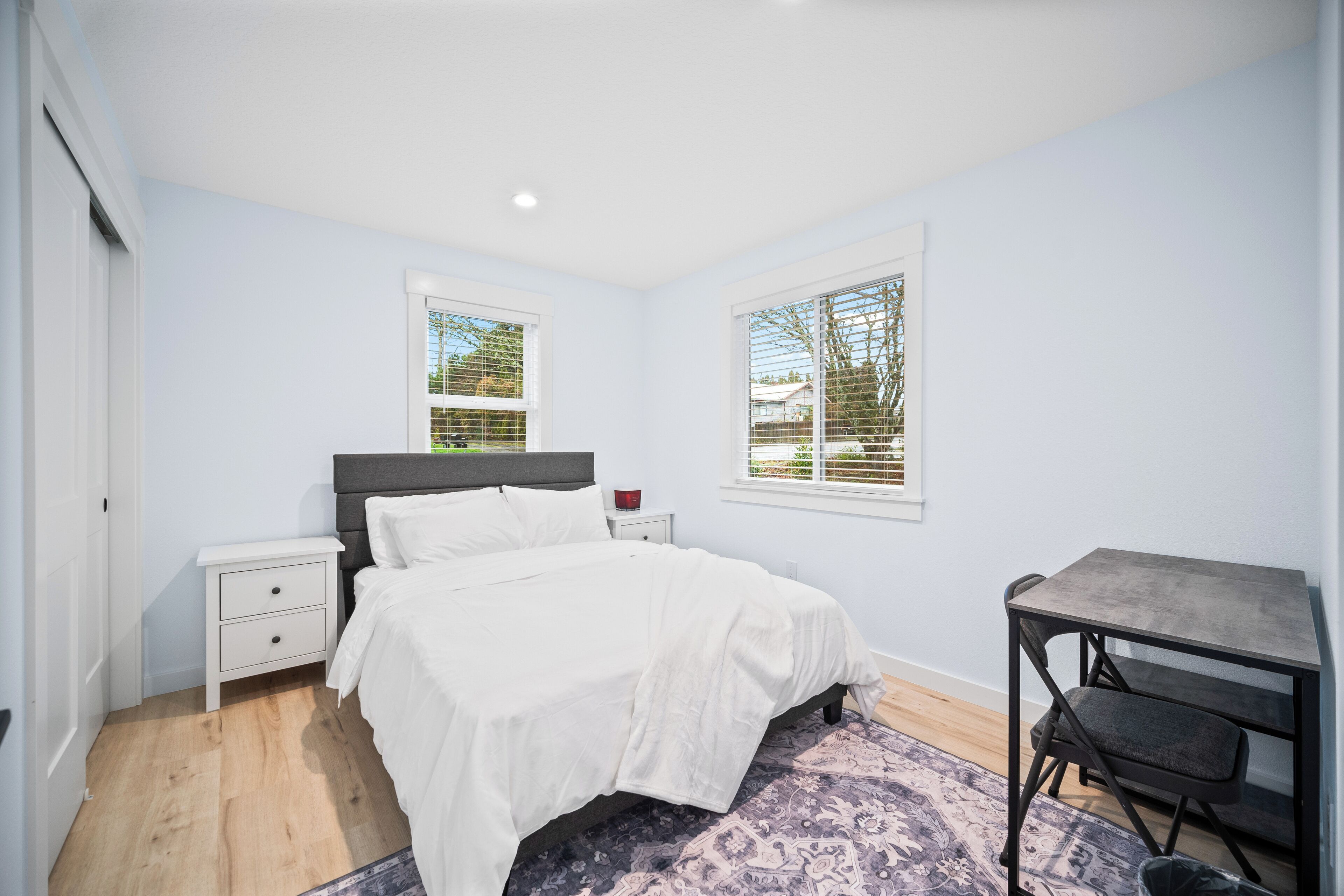 Quarto com cama, mesa de cabeceira, escrivaninha e janelas.