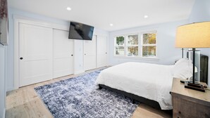 Een slaapkamer met een bed, een aan de muur bevestigde televisie, een raam met uitzicht op bomen en een lamp op een nachtkastje.