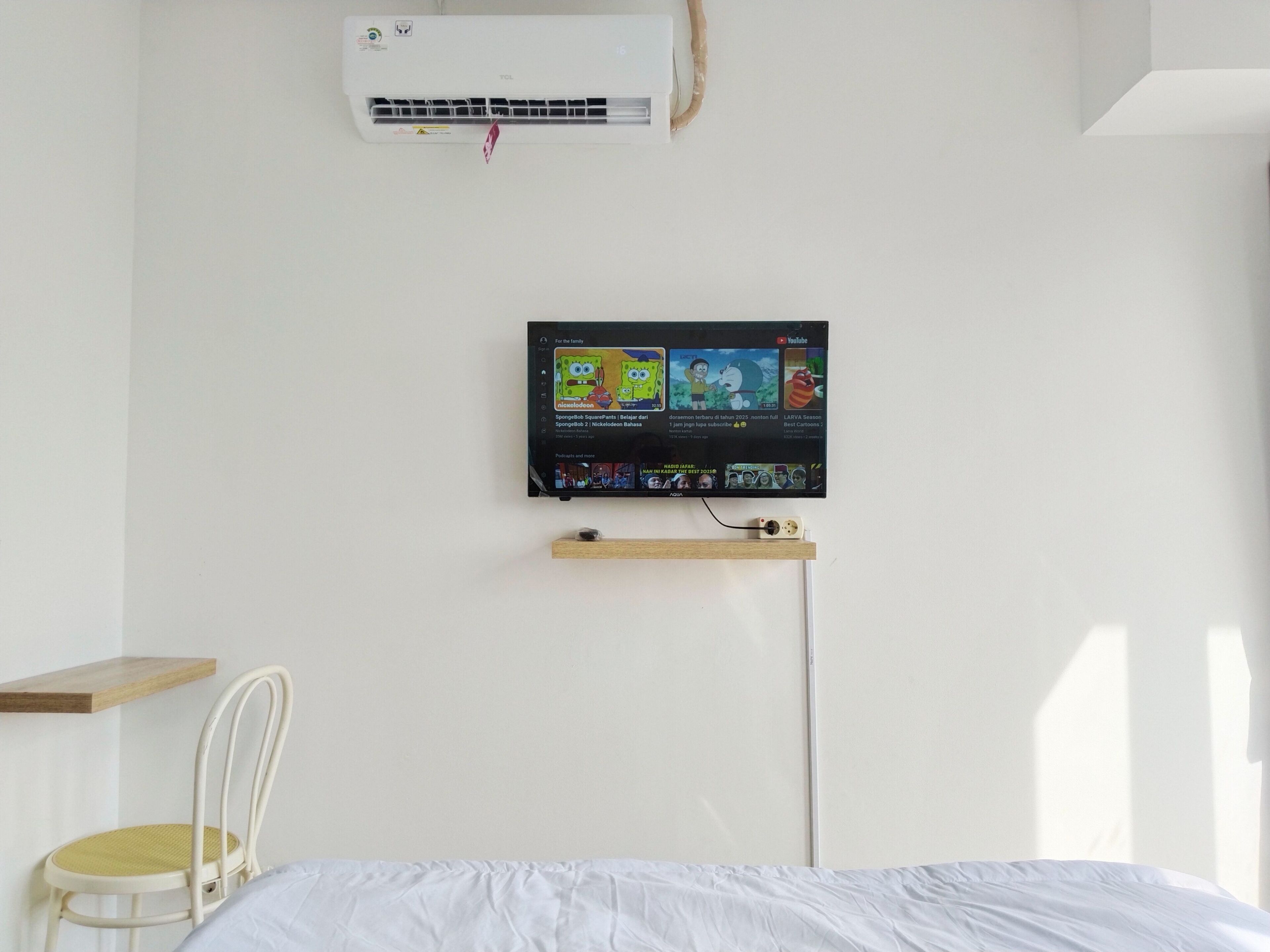 Ein Zimmer mit einem Bett, einer weißen Klimaanlage, einem an der Wand montierten Fernseher, der einen YouTube-Kanal anzeigt, einem Holzregal und einem weißen Stuhl.