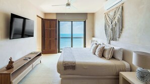 Un dormitorio con cama, televisor, ventilador de techo, ventana grande con vista al mar y aire acondicionado instalado en la pared.