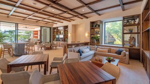 Amplia sala de estar con techo de madera, un conjunto de sofás, una mesa de comedor y una cocina con estantes y un ventanal.