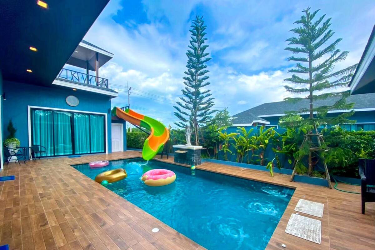 Une piscine avec un toboggan, entourée de plantes tropicales et dotée d’une terrasse en bois.