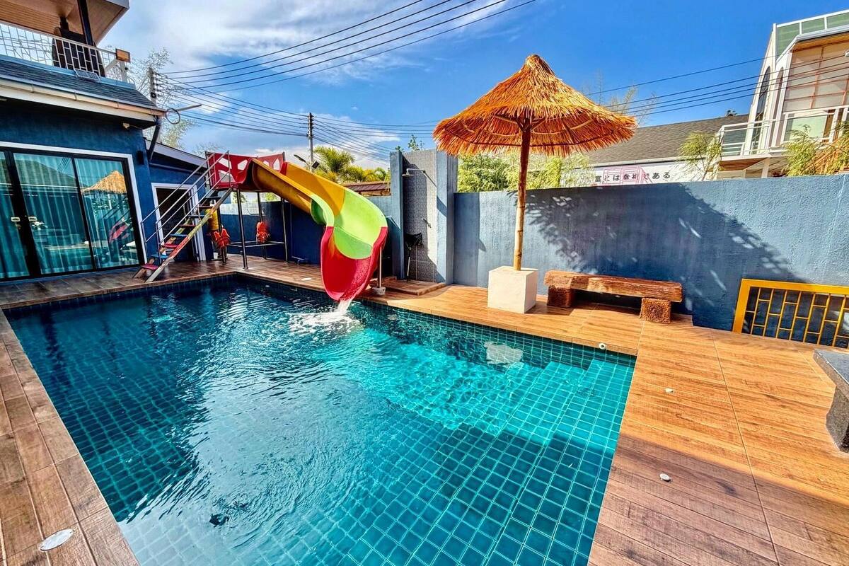 Une piscine avec un toboggan, une terrasse en bois et un parasol en chaume.