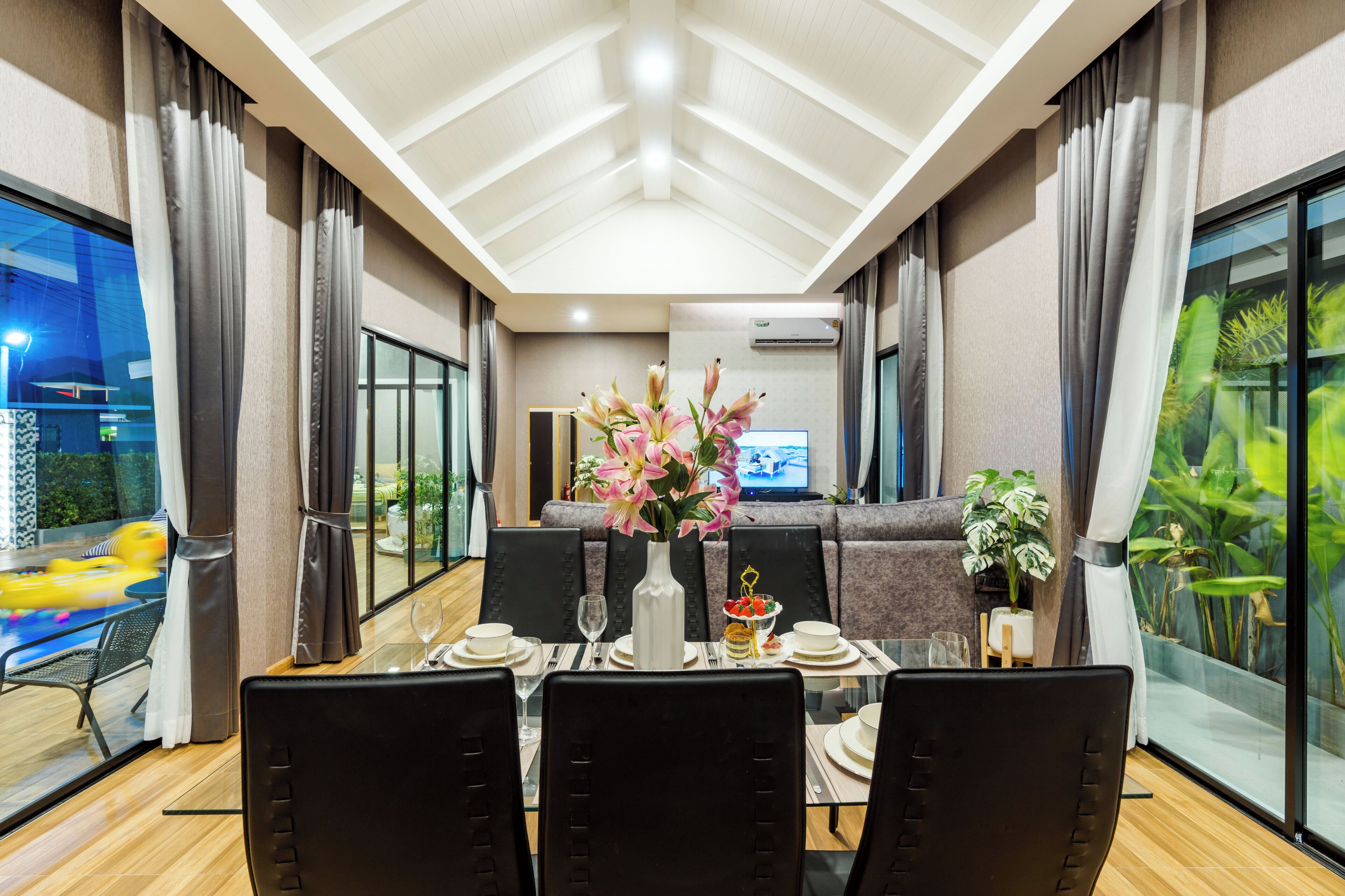 A dining area with a table set for four, a vase of flowers, and a view of a swimming pool and greenery outside.
