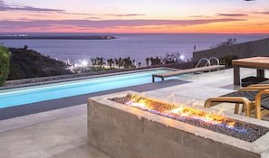 Una piscina con fogata, sillas de descanso y vistas al océano al atardecer.