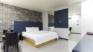 Ein modernes Hotelzimmer mit einer ziegelartigen Wand, einem großen Bett mit weißer Bettwäsche, einem dunkelblauen Kleiderschrank und einem kleinen Schreibtisch mit Stuhl.