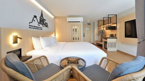 Habitación de hotel con una cama, dos sillas de mimbre, una mesita, televisión, kitchenette y aire acondicionado.