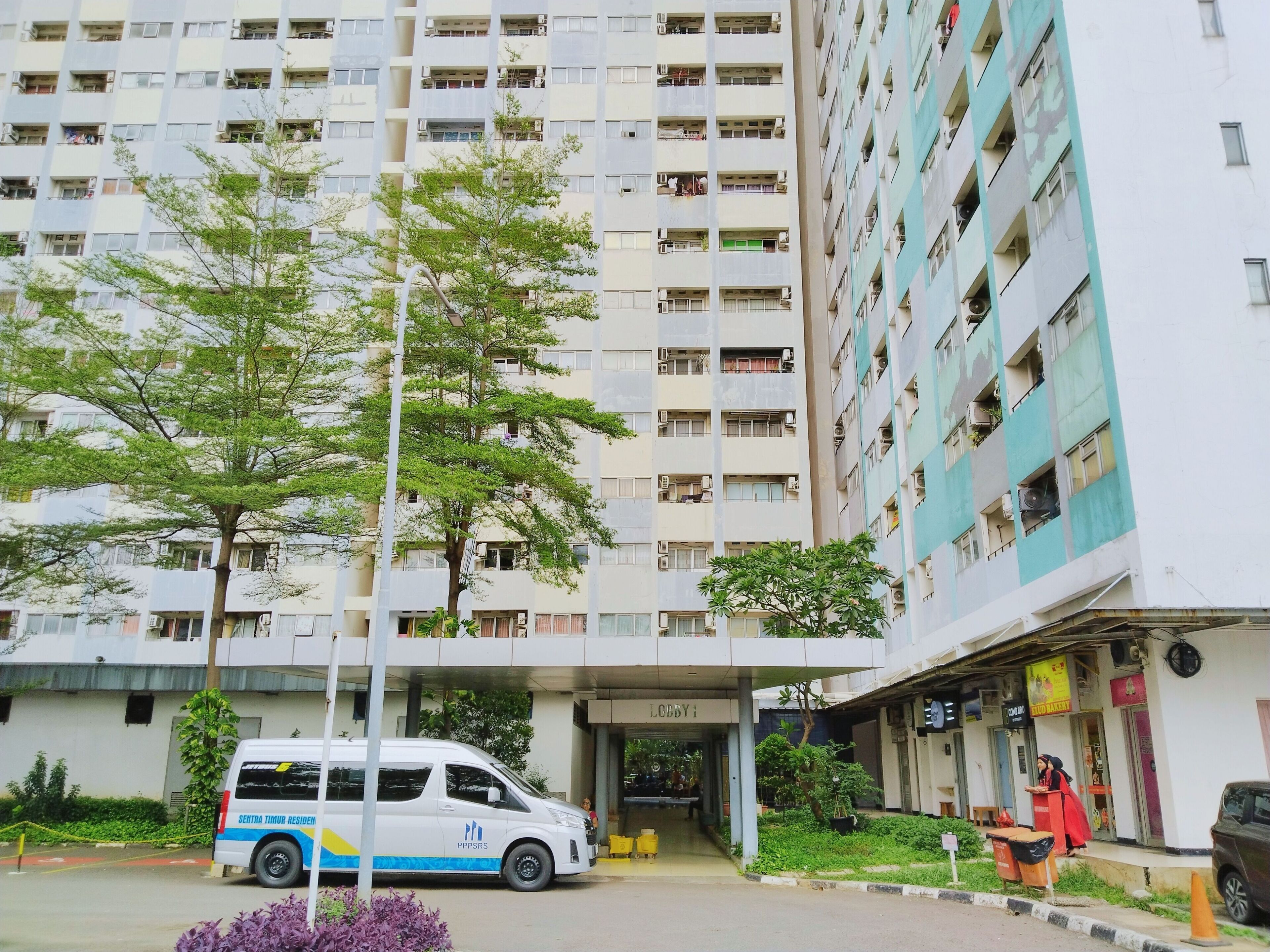 Un edificio residencial de varios pisos con fachada blanca y balcones. Una camioneta blanca con detalles azules y amarillos está estacionada en la parte delantera.