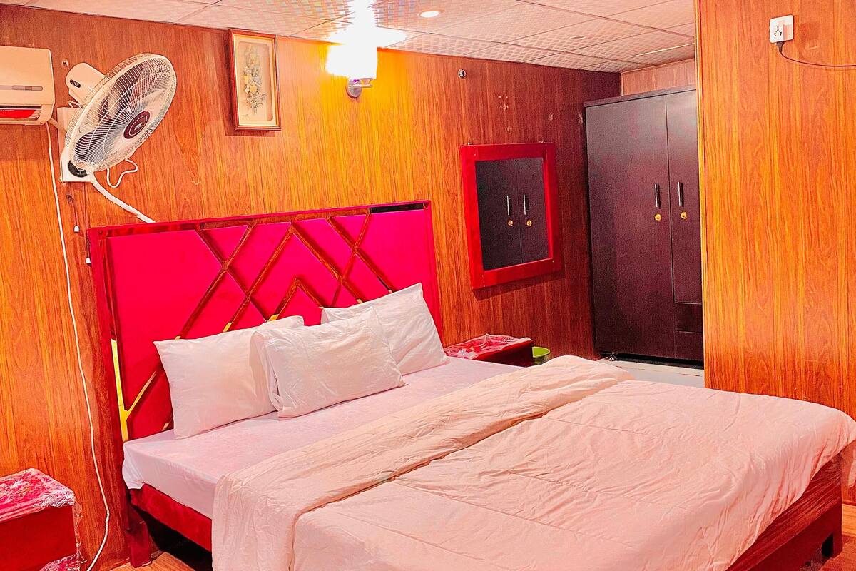 A hotel room with a bed, a red headboard, a wardrobe, a bedside table, a fan, and an air conditioning unit.