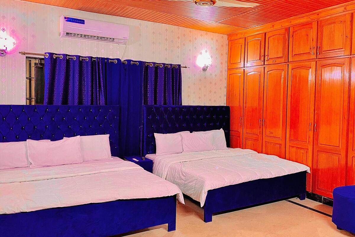 A room with two beds, a blue chair, wooden wardrobe, and an air conditioning unit.