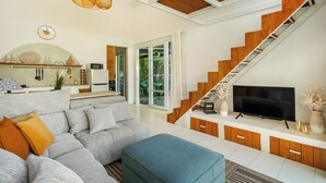 A modern living room with a grey sectional sofa, a blue ottoman, a wooden staircase, and a flat-screen TV. - Tropical 1BR Private Pool Villa, 8 mins to beach In Uluwatu (Pecatu)