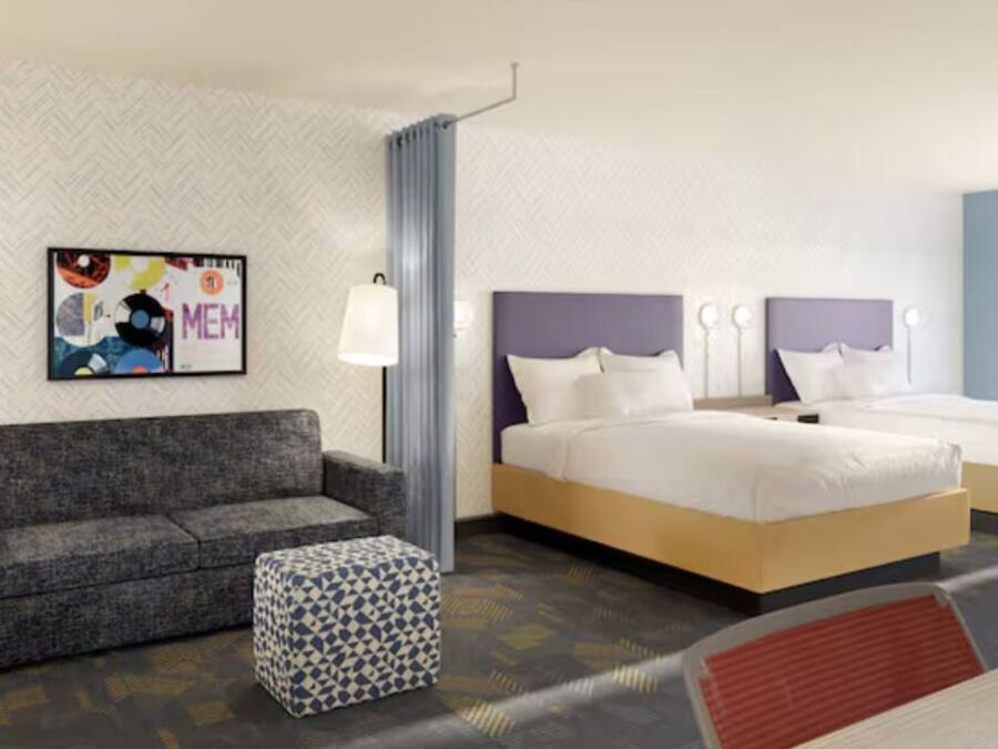 Habitación de hotel con un sofá gris, dos camas individuales, un reposapiés estampado, una lámpara fijada a la pared y una obra de arte enmarcada.