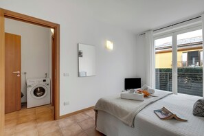 A bedroom with a washing machine, a bed with a blanket, a bedside table with a book, and a window with a view of a building. - Residence Due Laghi (Ispra)
