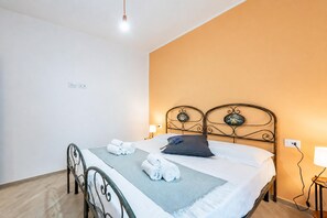 Un lit bien fait, recouvert d’une couverture grise et agrémenté d’un seul oreiller foncé, deux serviettes enroulées posées sur le lit, une tête de lit en métal et une lampe de chevet.