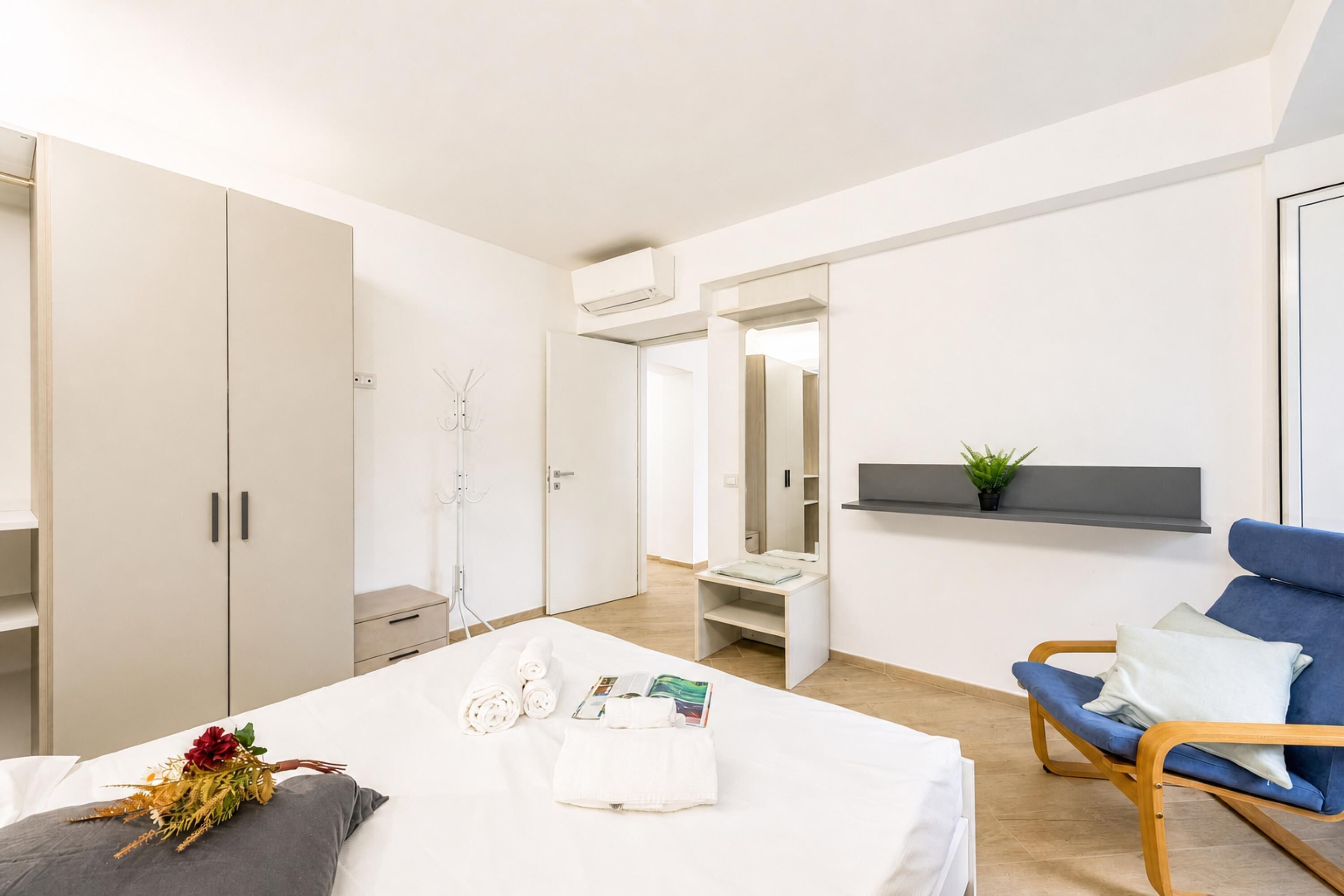 A modern hotel room with a bed, a blue chair, a wardrobe, and a small plant on a shelf.