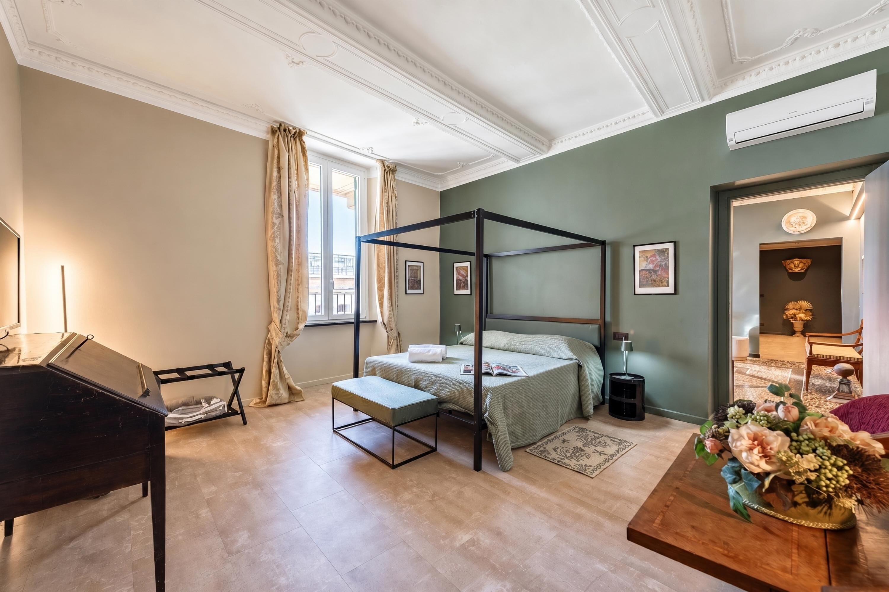 A bedroom with a four-poster bed, a piano, a wooden coffee table, and a floral arrangement.