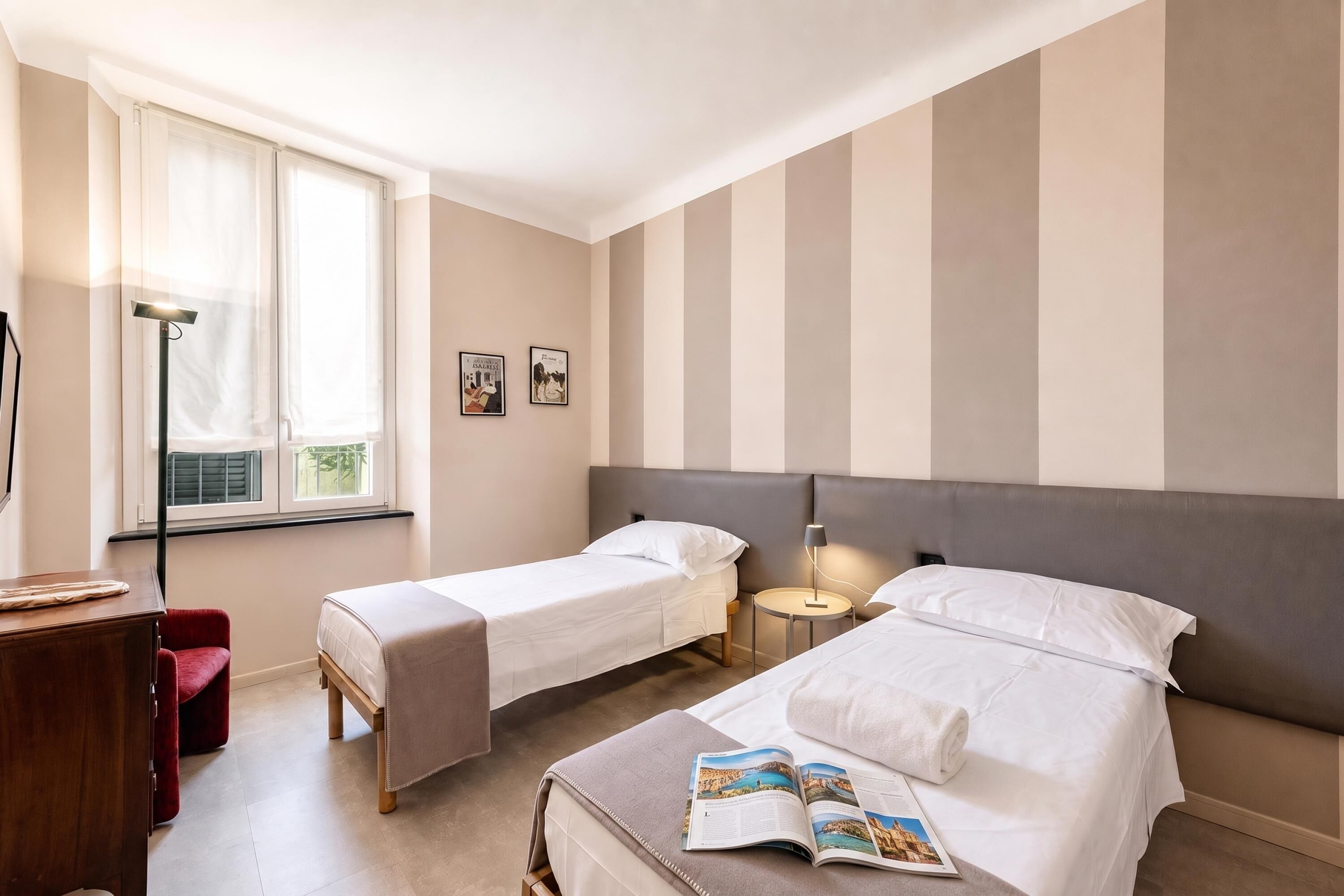 A hotel room with two single beds, a desk, a chair, a lamp, and a window with curtains.