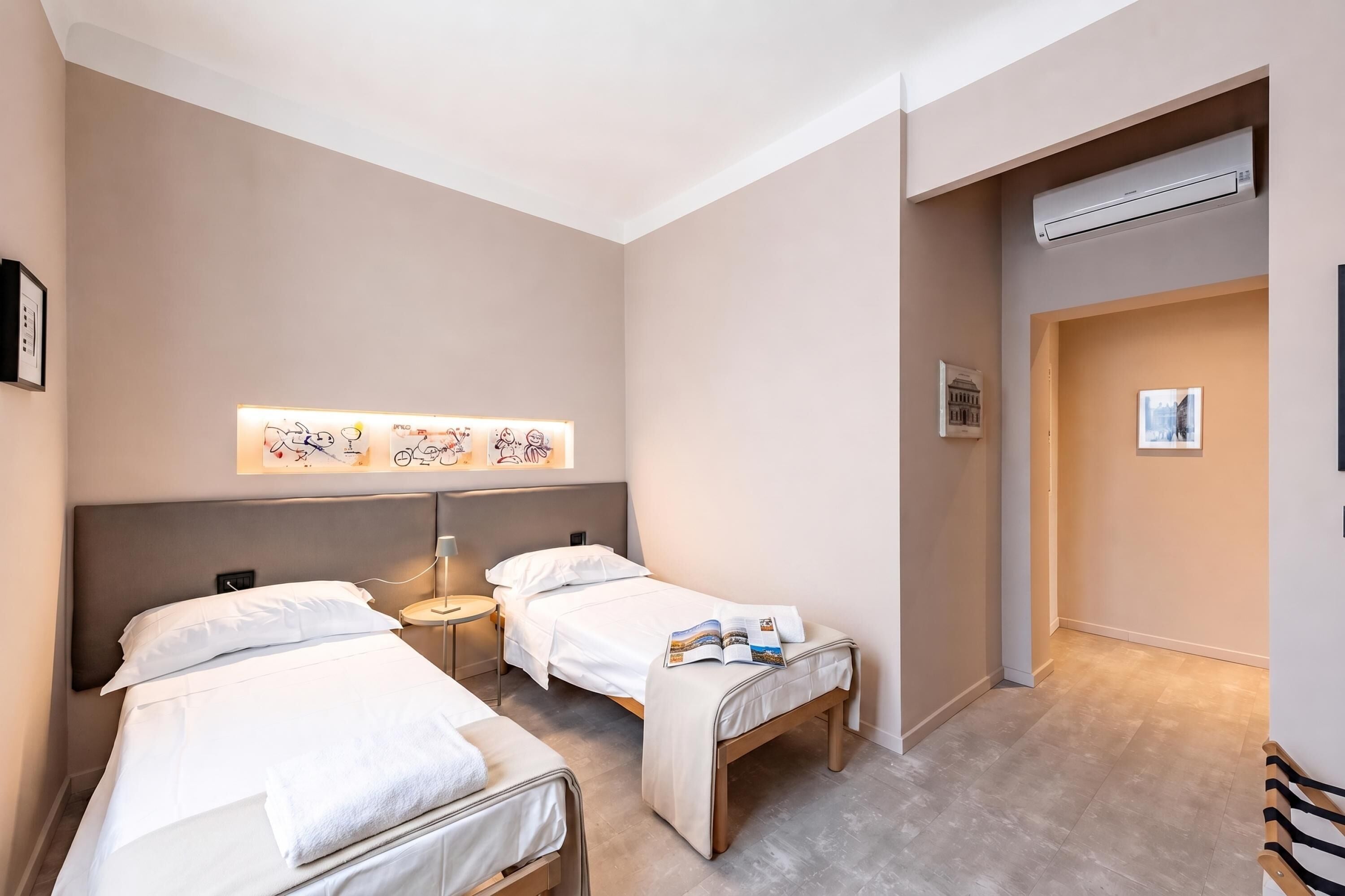 A hotel room with two beds, a bedside table with a lamp, a wall-mounted air conditioner, and a framed picture on the wall.