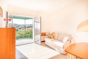 A modern living room with a white sofa, a wooden table, and a glass door leading to a balcony with a city view.