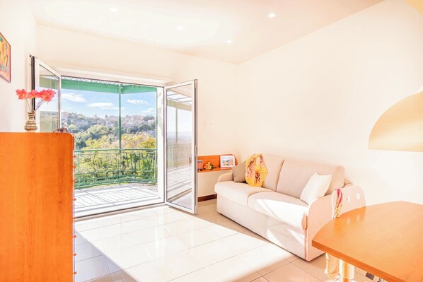 A bright living room with a white sofa, a wooden table, and a sliding glass door leading to a balcony with a city view.