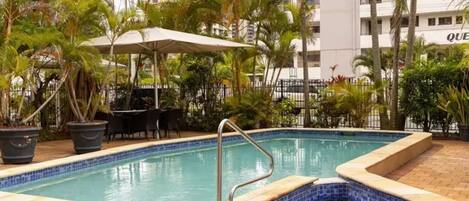 A swimming pool with a tiled edge, surrounded by palm trees and a patio area with outdoor furniture.